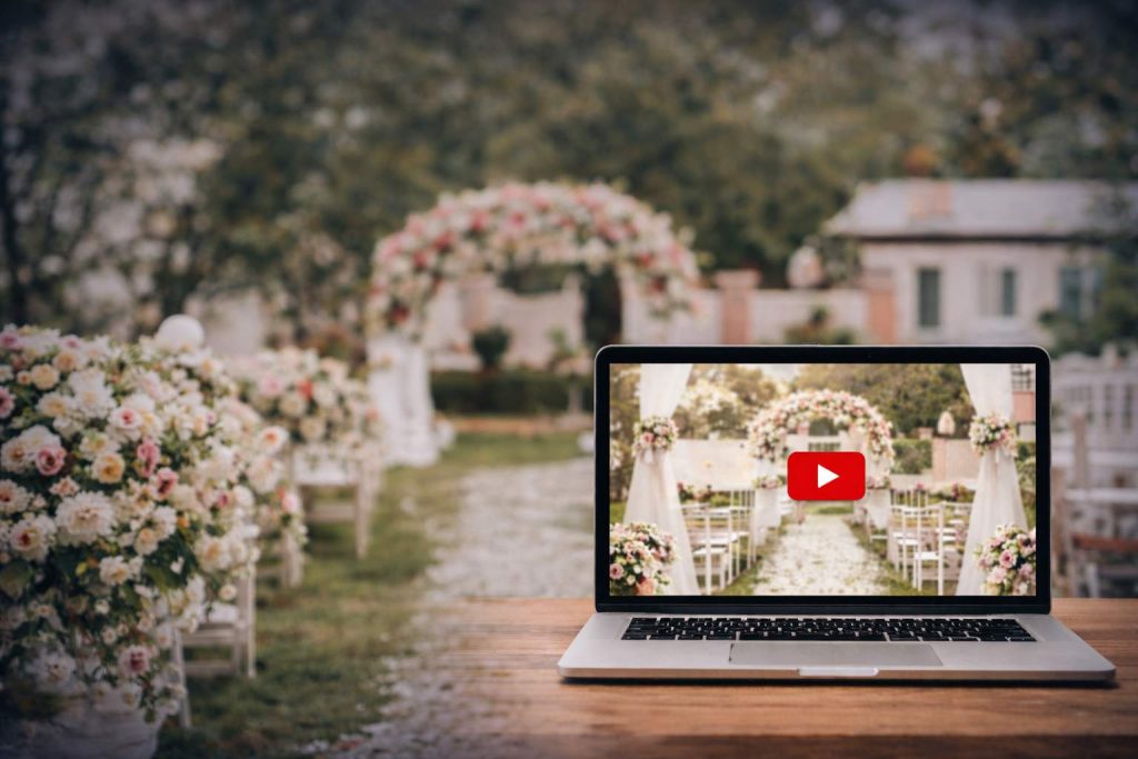laptop with YouTube video ad logo on the screen sitting on a wooden table with an outdoor wedding setup with white and pink flower stands behind the chairs